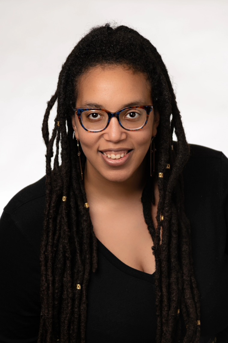 African American woman wearing glasses with long locs, wearing a black  shirt sitting against a white background