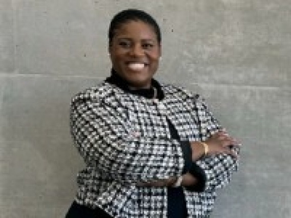 African MAerican woman stand in font of a grey congrete wall. She is wearing a black and white jacket and black dress. She is smiling with her arms folded across her chest