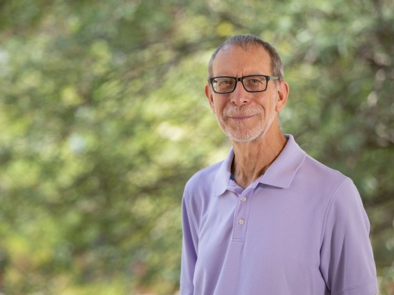 White male stand in front of blurry greenery background. He has grey hair and is wearing glasses and a lavender polo
