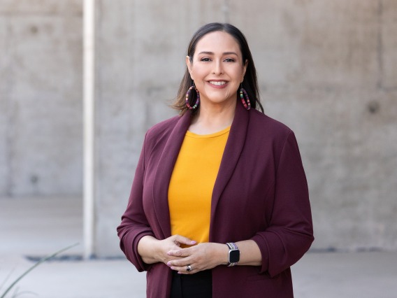 Esperanza is standing in from of a concrete structure. She is wearing a burgandy blazer, mustard color top, and black pants. She is smiling with her hand slightly touching at the wait. 