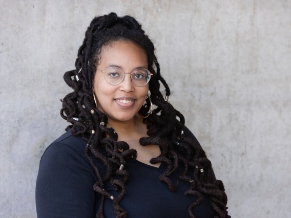 Val is infront of a concrete wall. She is wearing a black top. Her locs are half up half down with gold embellishments throughout and curly. She is wearing glasses and smiling. 