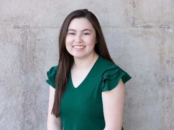 CeCe is in front of a  concrete wall. She is wearing a dark green blouse. She has dark brown hair that is parted to the side and down and she is smiling. 