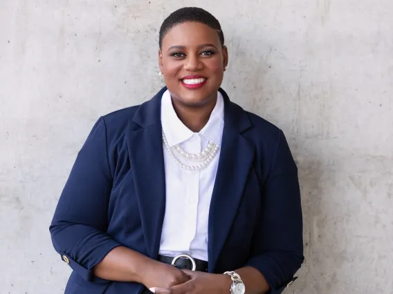 Sabrina is leaning against a concrete wall wearing a navy blue blazer, White shirt, and black pants. She has her finger interlocked at the waist and is smiling