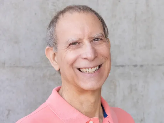 Roy is in front of a concrete wall. He is wearing a peach colored blazer and smiling. 