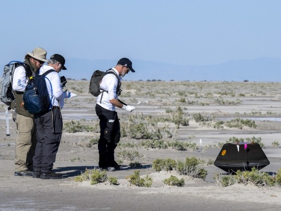 three people approaching a tire-sized capsule in the middle of a desert landscape