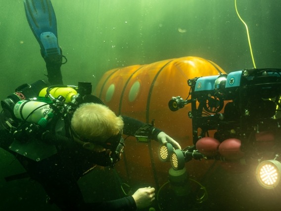 A scuba diver services an underwater research robot with an underwater camping tent in the background, located at the University of Arizona&#039;s Biosphere 2.