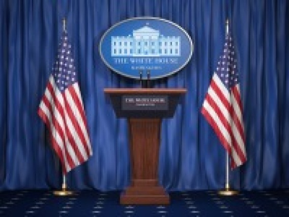 A podium stands before a blue background, between a pair of american flags. A White House emblem can be seen behind the podium.