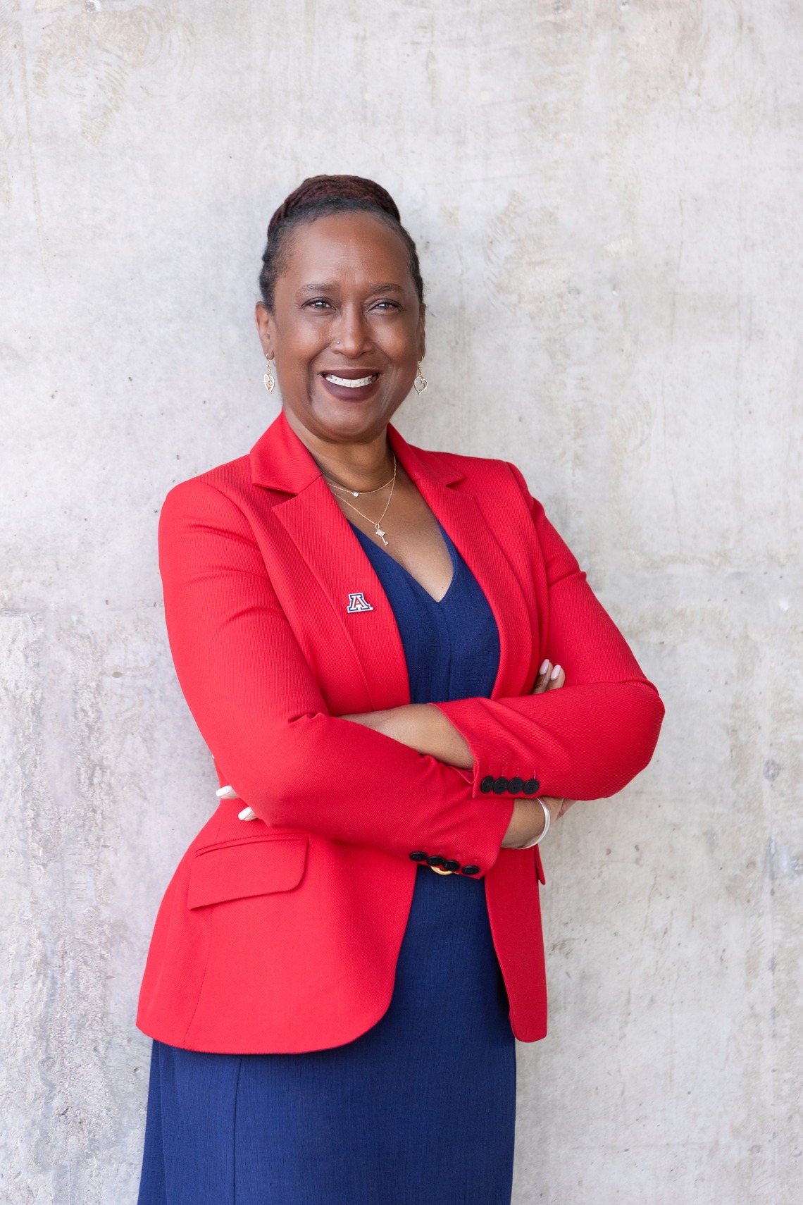 Dr. Hatcher is wearing a red blazer with a University Of Arizona "A" Pin and a dark blue dress. She is smiling with her arms crossed as she stands against a concrete wall. 