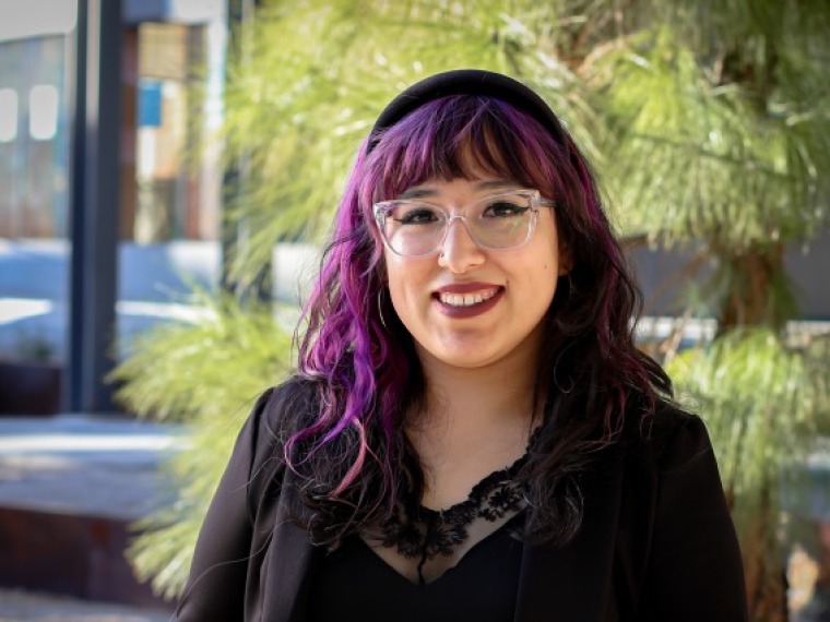 Against the background of trees, women with black and purple hair smiles. She is wearing a black button shirt and glasses
