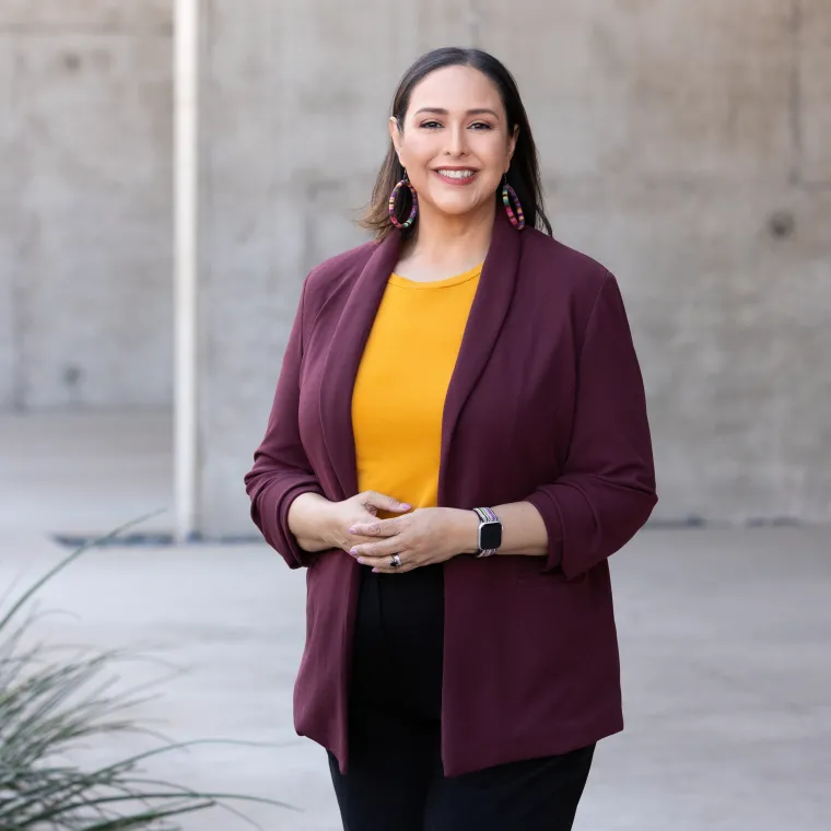 Esperanza is standing in from of a concrete structure. She is wearing a burgandy blazer, mustard color top, and black pants. She is smiling with her hand slightly touching at the wait. 