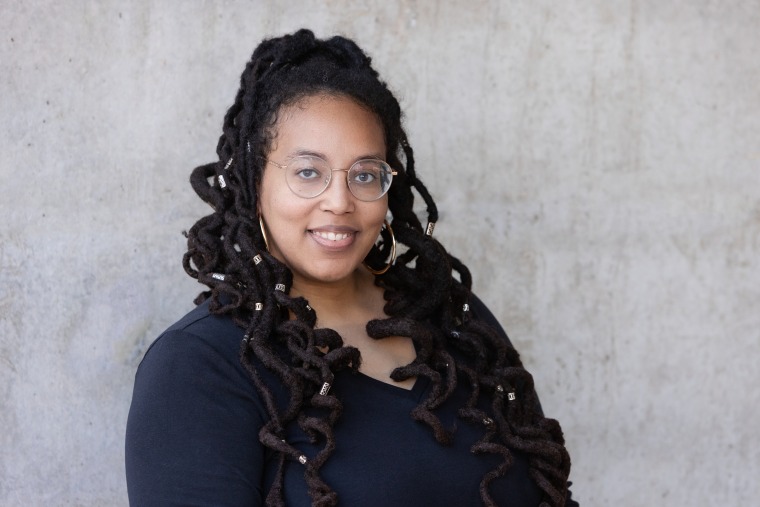 Val is infront of a concrete wall. She is wearing a black top. Her locs are half up half down and curly. She is wearing glasses and smiling. 