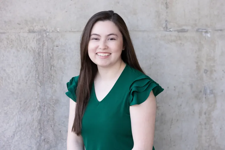 CeCe is in front of a  concrete wall. She is wearing a dark green blouse. She has dark brown hair that is parted to the side and down and she is smiling. 