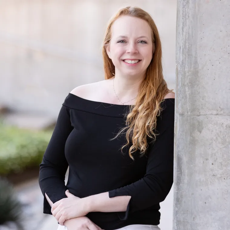 Joelle is leaning against a concrete wall with one hand over her wrist. She is smiling wearing a black shirt and light colored pants.