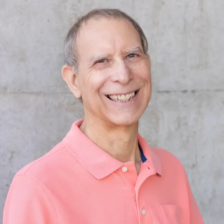 Roy is in front of a concrete wall. He is wearing a peach colored blazer and smiling. 