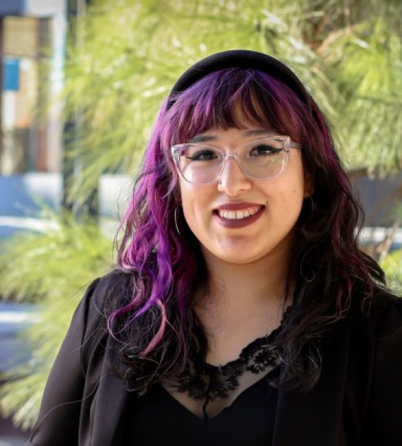 Against the background of trees, women with black and purple hair smiles. She is wearing a black button shirt and glasses
