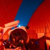 lisa garcia looking through a telescope under a dome lit with red light slightly open to show a starry sky